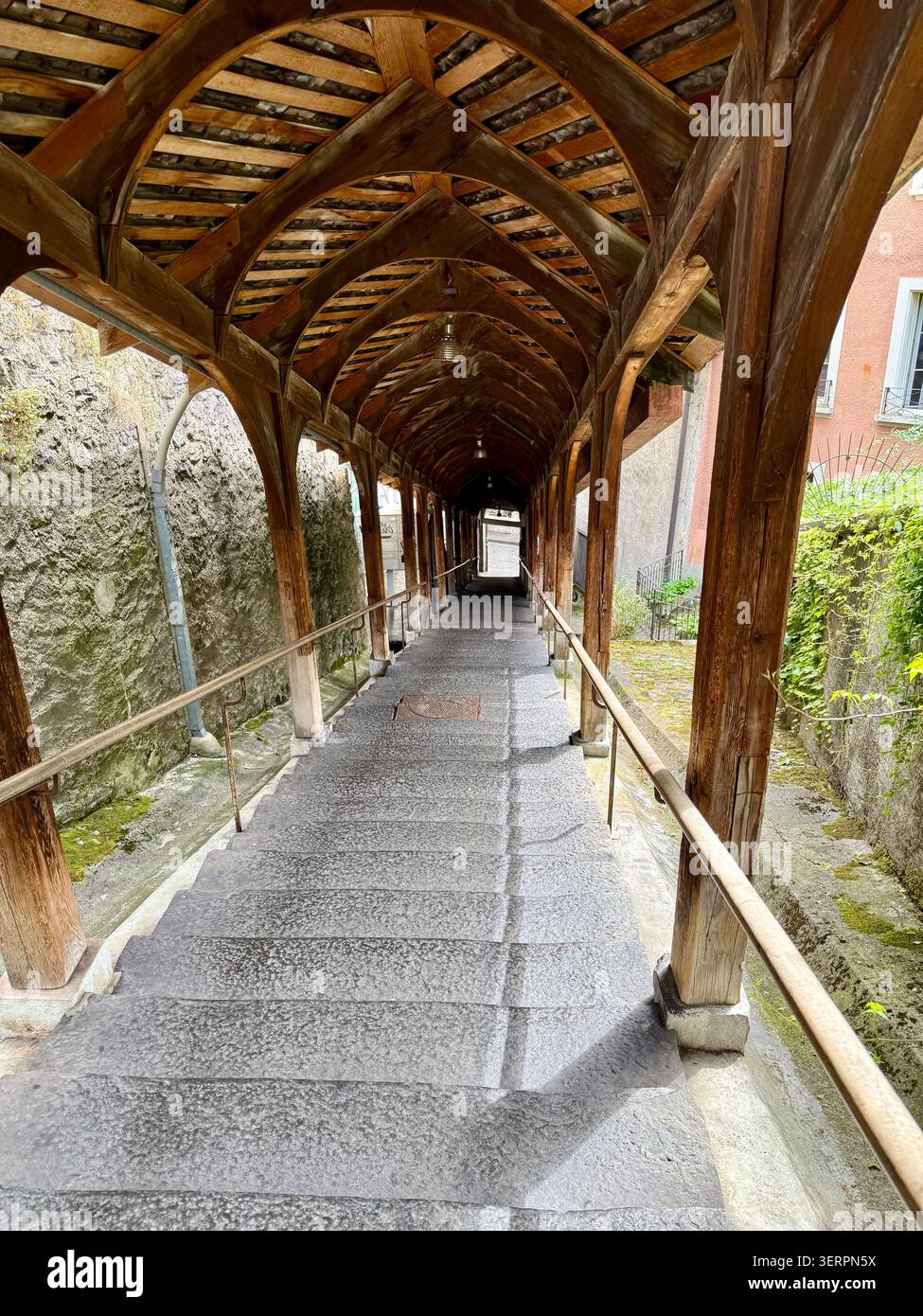 Una storica scalinata coperta in legno con travi ad arco in legno e gradini in pietra usurata conduce attraverso un passaggio simile a un tunnel, fiancheggiato da una wal in pietra intemprata - Immagine stock catturata con smartphone