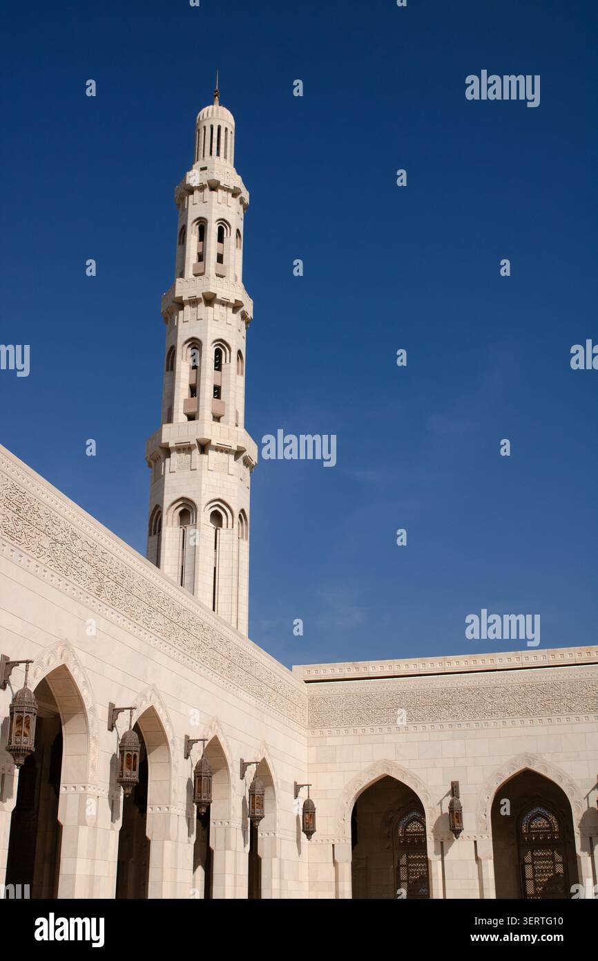 Vista dell'alto minareto e dei passaggi ad arco con lanterne appese alla grande Moschea del Sultano Qaboos sotto un cielo azzurro a Mascate, governatore di Mascate Foto Stock