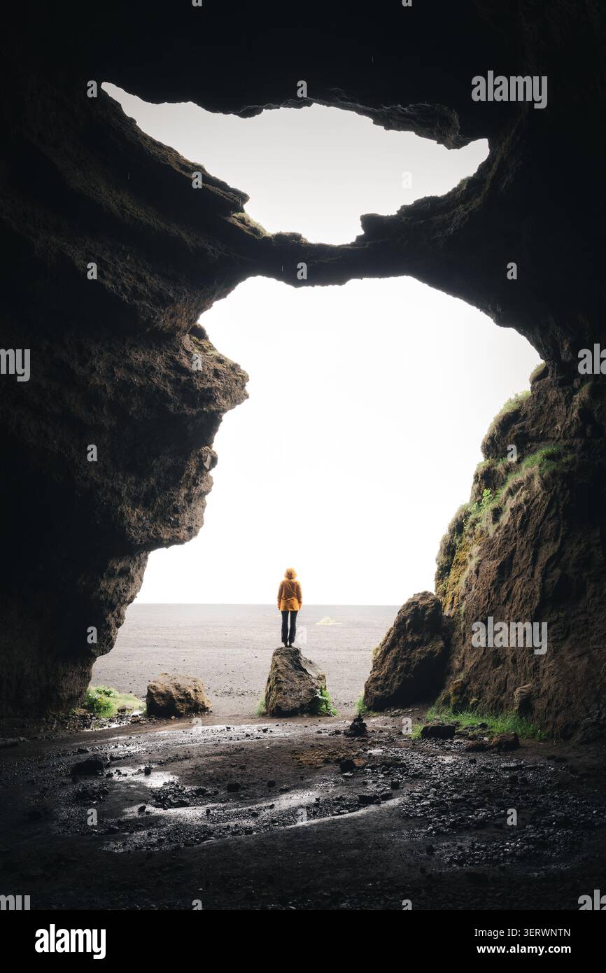 Vista di una persona in una giacca gialla in piedi su una roccia all'interno di una buia grotta vulcanica Yoda Caveopening che guarda verso un luminoso paesaggio coperto IC Foto Stock
