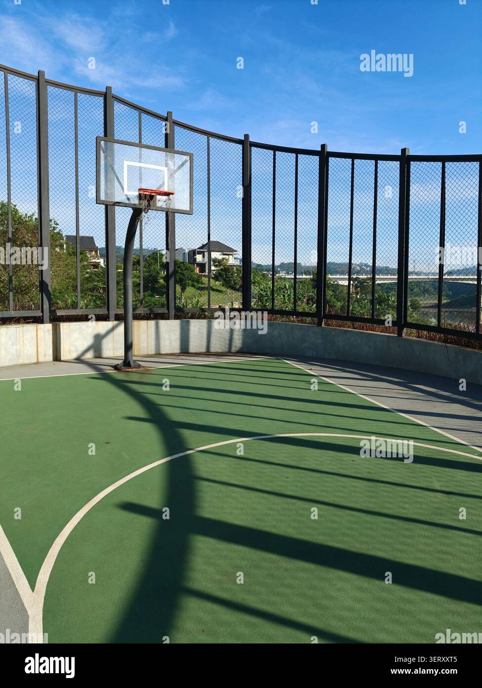 Campo da basket all'aperto con superficie verde, canestro, tabellone e recinzione protettiva alta sotto un cielo azzurro limpido in una giornata soleggiata - Immagine stock catturata con smartphone
