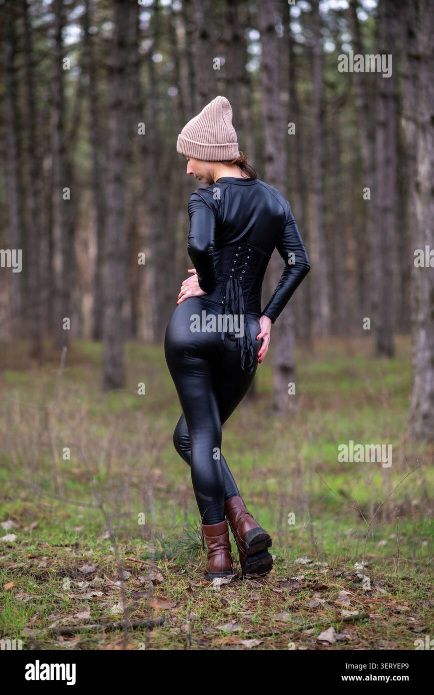 Ritratto posteriore di una donna che indossa un abito e un berretto in ecopelle nera, che cammina attraverso una pineta con stivali marroni. Foto Stock