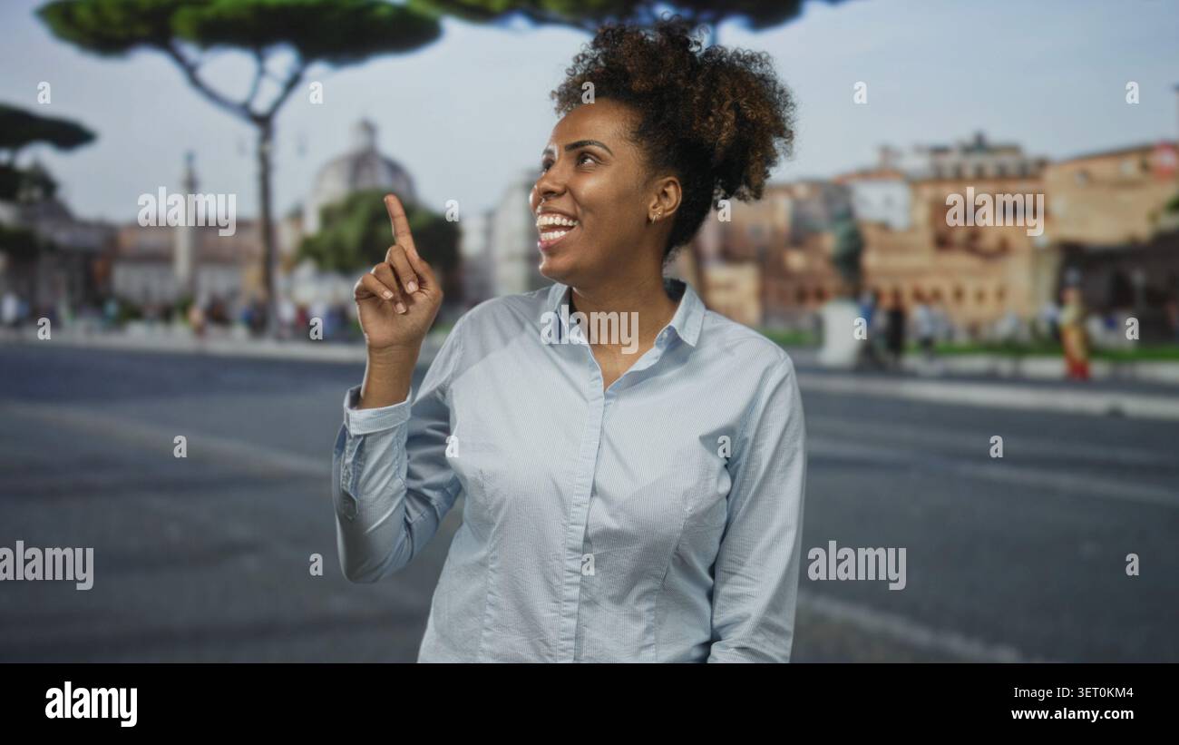 Donna con i capelli ricci punta il dito verso l'alto e sorride in grande mentre si trova in una strada della città costeggiata da edifici storici e pedoni; curiosità Foto Stock