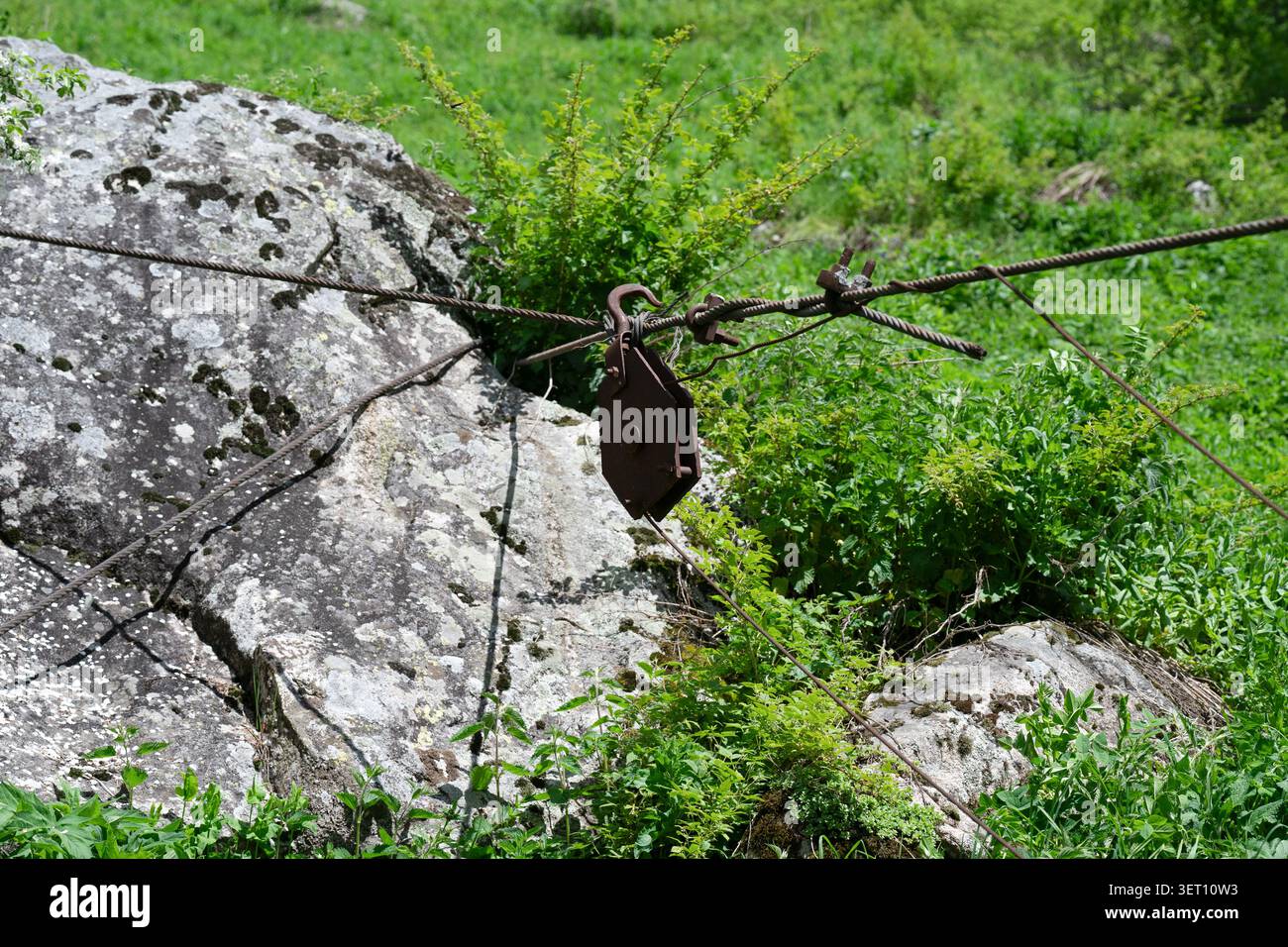 Blocco di ferro vintage con funi metalliche su uno sfondo di erba verde. Vecchio blocco puleggia arrugginito e cavi in acciaio all'aperto vicino a un grande masso. Foto Stock