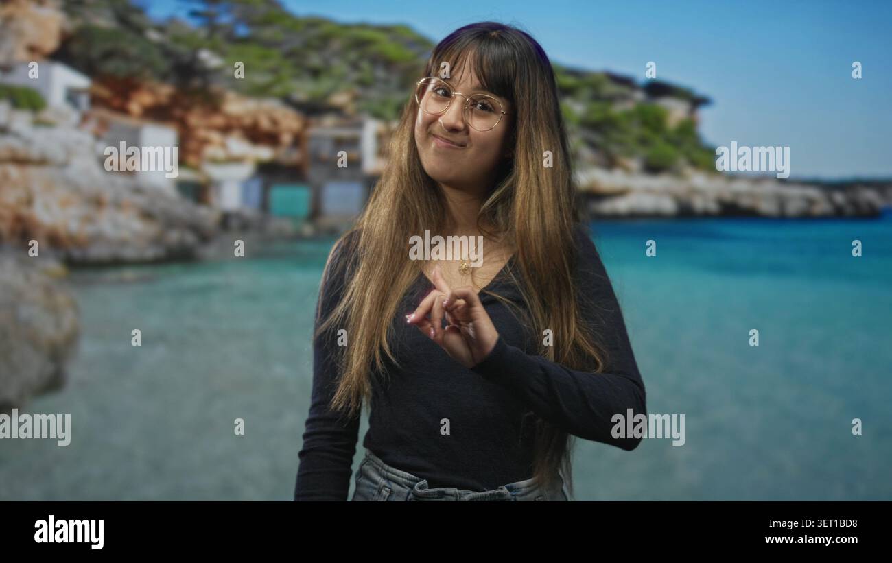 La giovane donna punta il dito indice verso la fotocamera sulla spiaggia, indossando occhiali e denim, capelli lunghi visibili, sorridendo delicatamente; giocosa sicurezza. Foto Stock