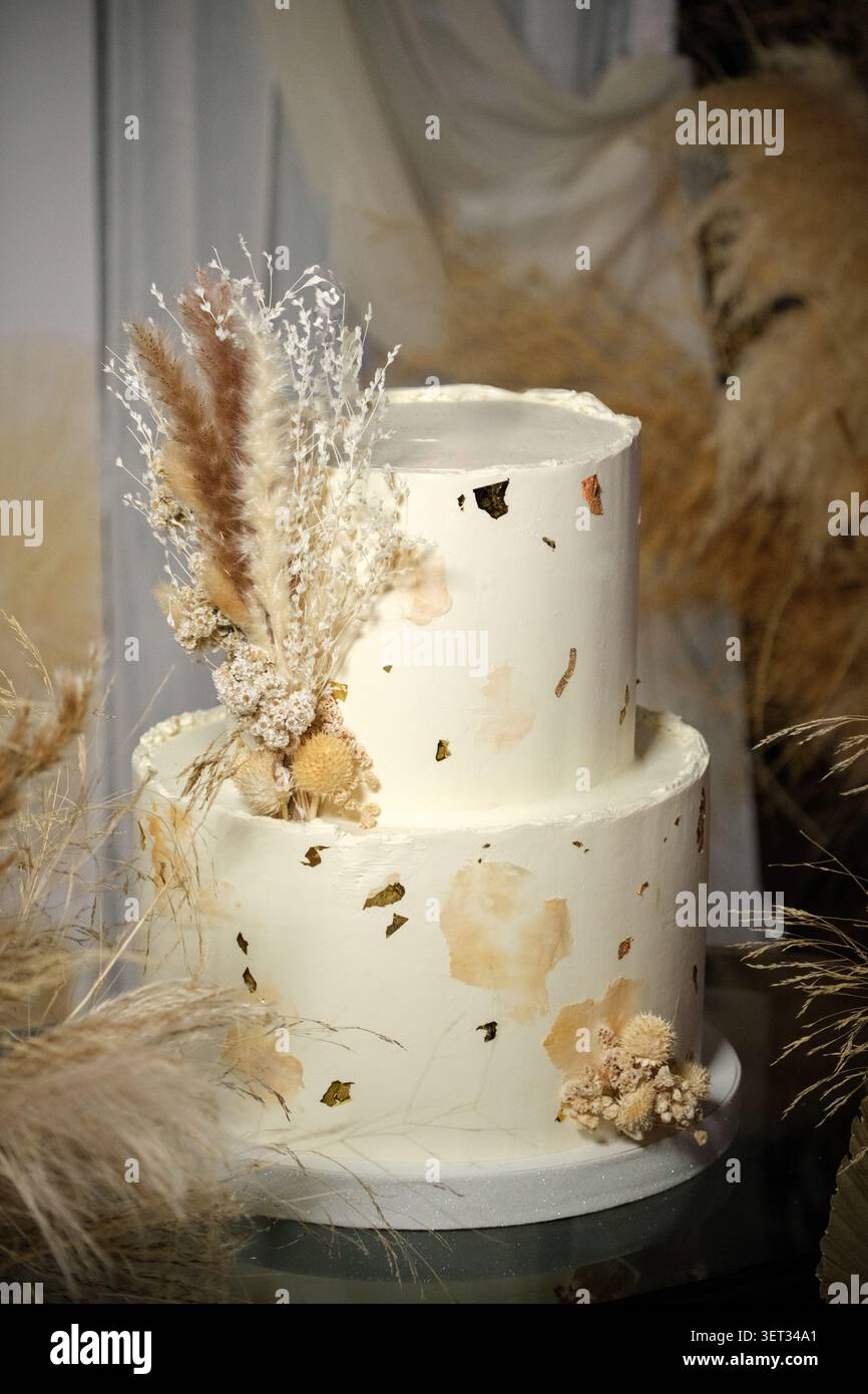 Un'elegante torta nuziale a due piani decorata con foglie d'oro e fiori secchi, esposta in un ambiente boho rustico con un'atmosfera morbida e romantica. Foto Stock