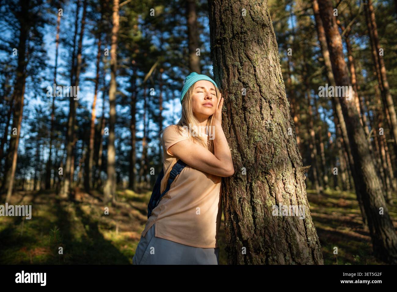 Donna rilassata che tocca il tronco dell'albero nella foresta, gli occhi chiusi e si crogiola alla luce del sole. Senza stress. Foto Stock
