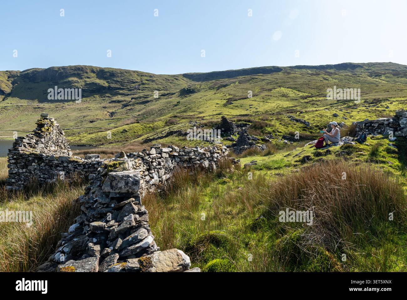 Donna che fa uno spuntino seduto tra le rovine del villaggio abbandonato di carestia di Lough Coolin degli anni '1840 in Irlanda Foto Stock