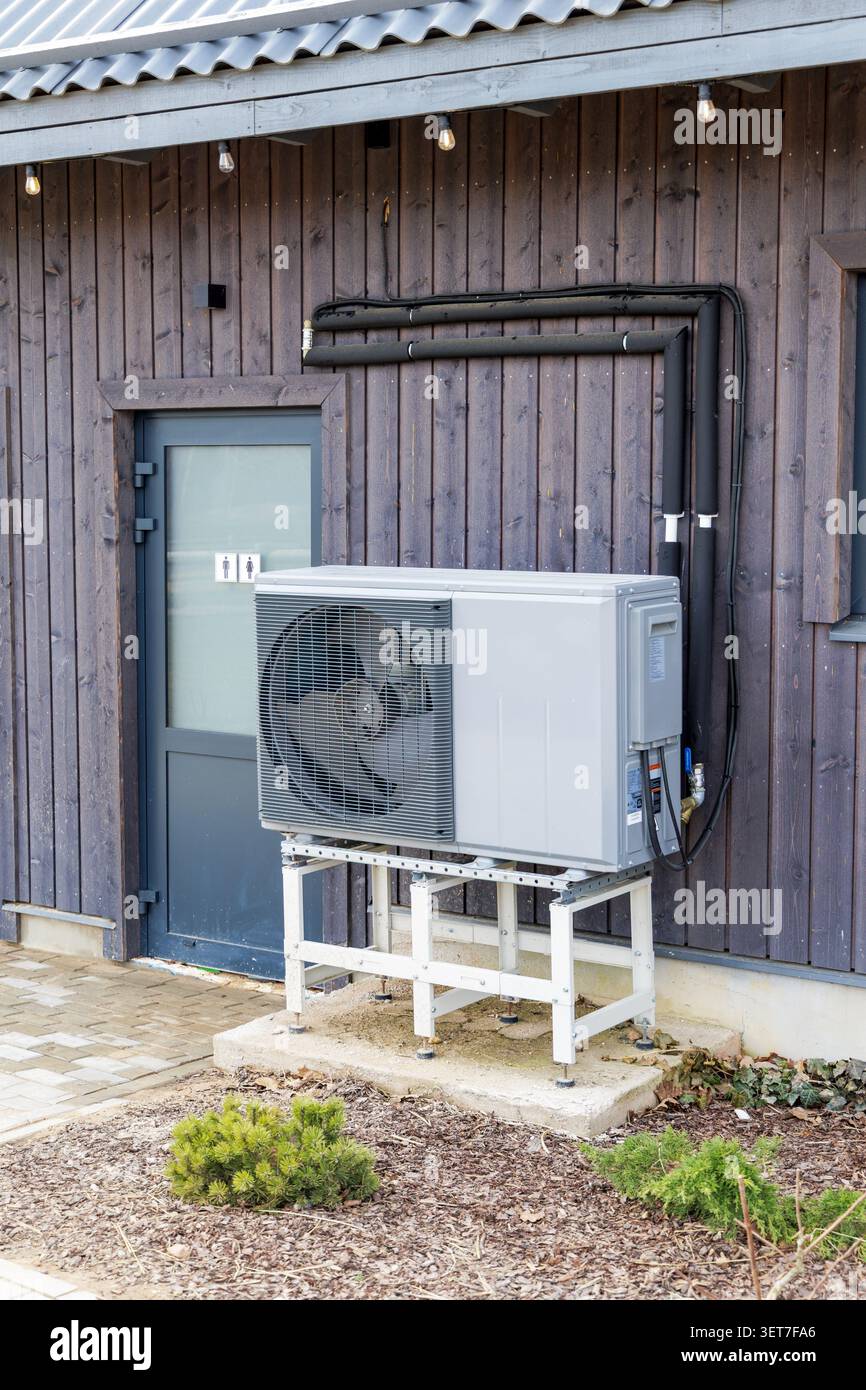 Unità esterna di un efficiente sistema a pompa di calore della fonte d'aria installata all'esterno di una casa di legno scuro accanto alla porta di un bagno pubblico. Foto Stock