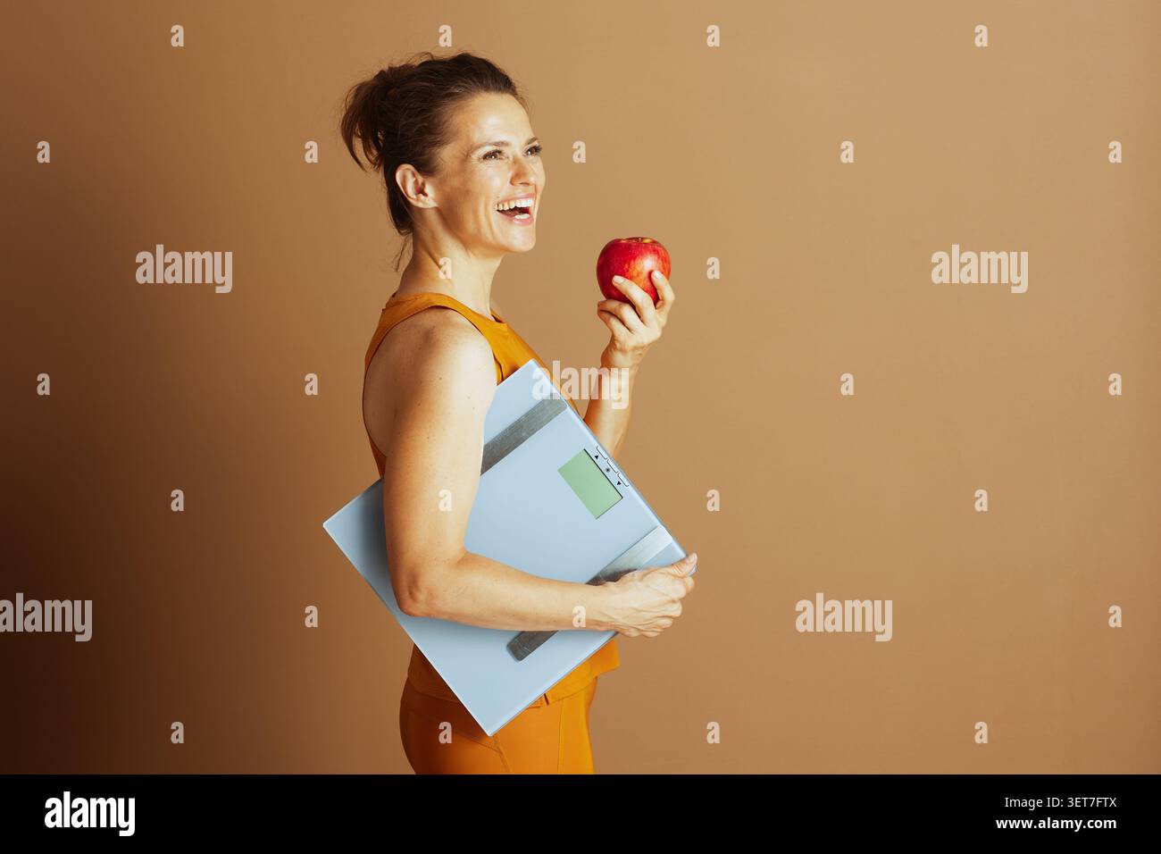Una donna sorridente di mezza età in abbigliamento sportivo arancione, mostrata nel profilo, ha una mela e una scala digitale, simboleggiando la salute, il fitness e una a motivata Foto Stock
