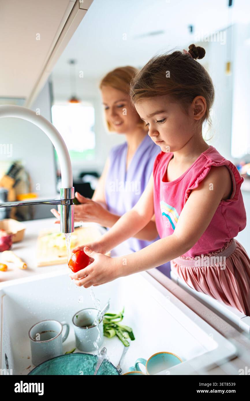 Una ragazza lava i pomodori mentre sua madre prepara il pranzo Foto Stock