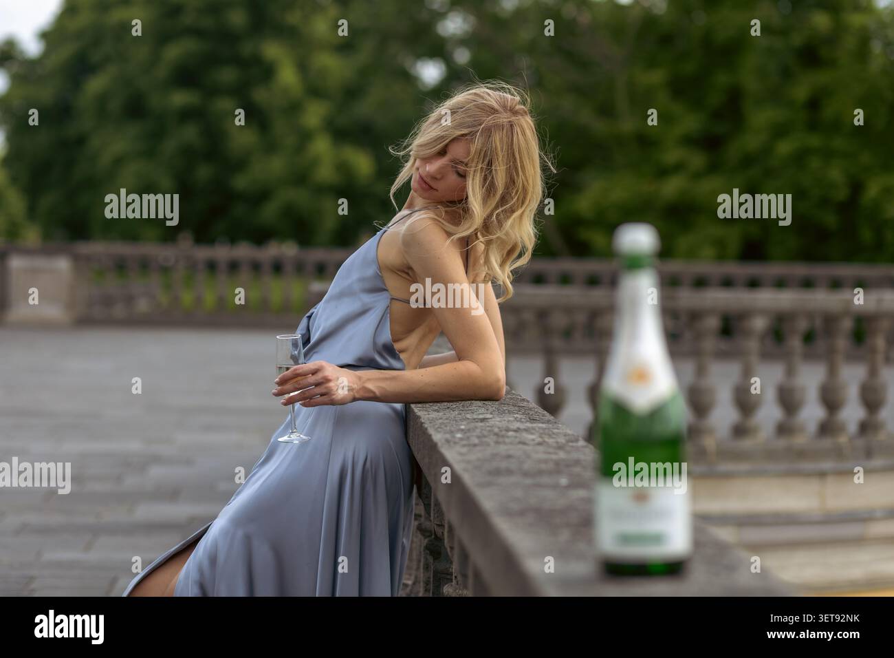 Una donna si appoggia su una ringhiera di pietra in un parco. Tiene un drink mentre guarda lontano. Gli alberi sono sullo sfondo sotto un cielo limpido. Foto Stock