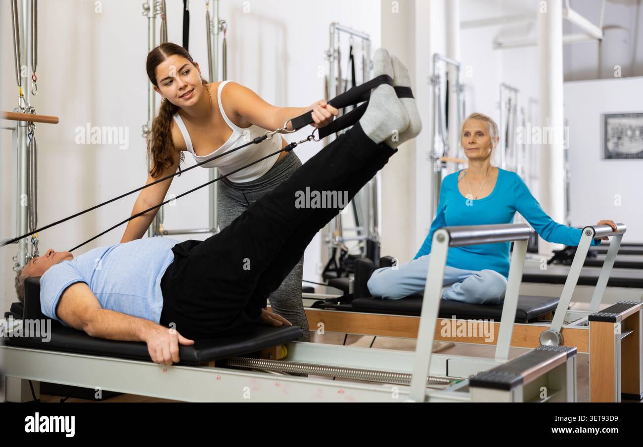 Giovane istruttrice positiva che controlla i movimenti dell'uomo anziano che si allunga sulle attrezzature del riformatore Pilates in studio Foto Stock