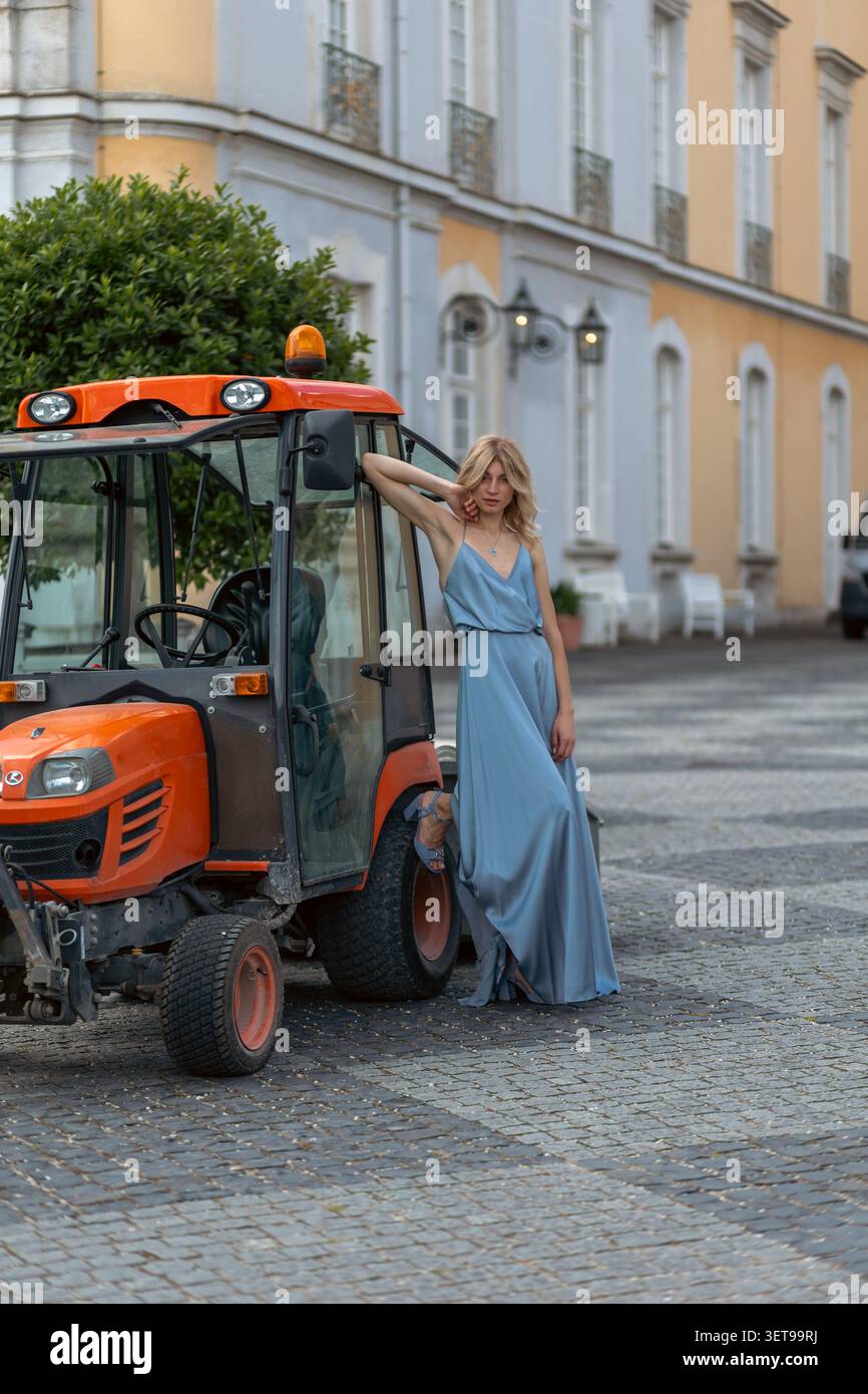 Una donna sta vicino a un trattore in una strada di città. Indossa un vestito blu e si appoggia al trattore. Gli edifici e un albero sono sullo sfondo durando Foto Stock
