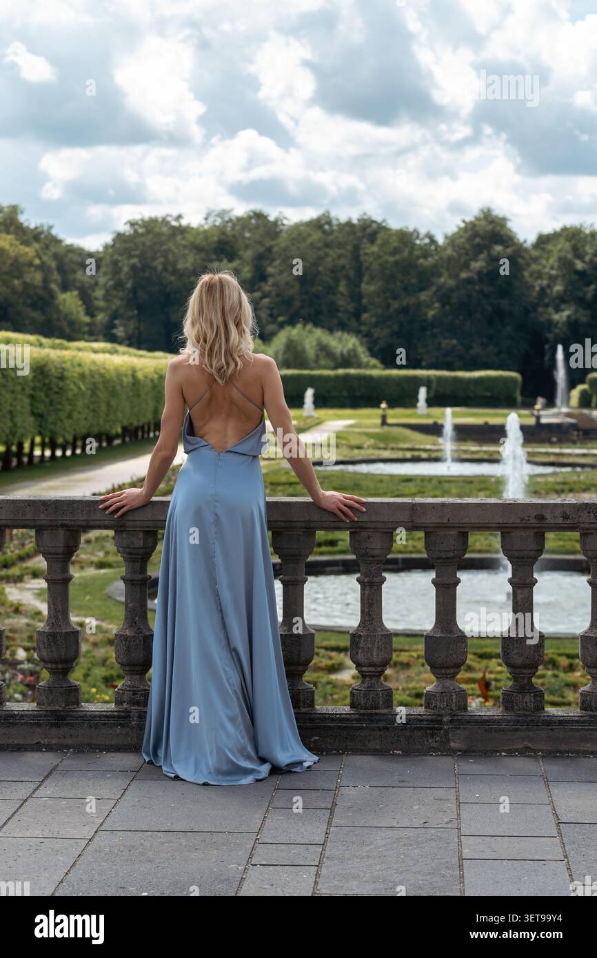 Una donna con un vestito leggero si appoggia contro una ringhiera di pietra. Si affaccia su un grande giardino con fontane, alberi e un cielo ampio. E' estate e l'ar Foto Stock