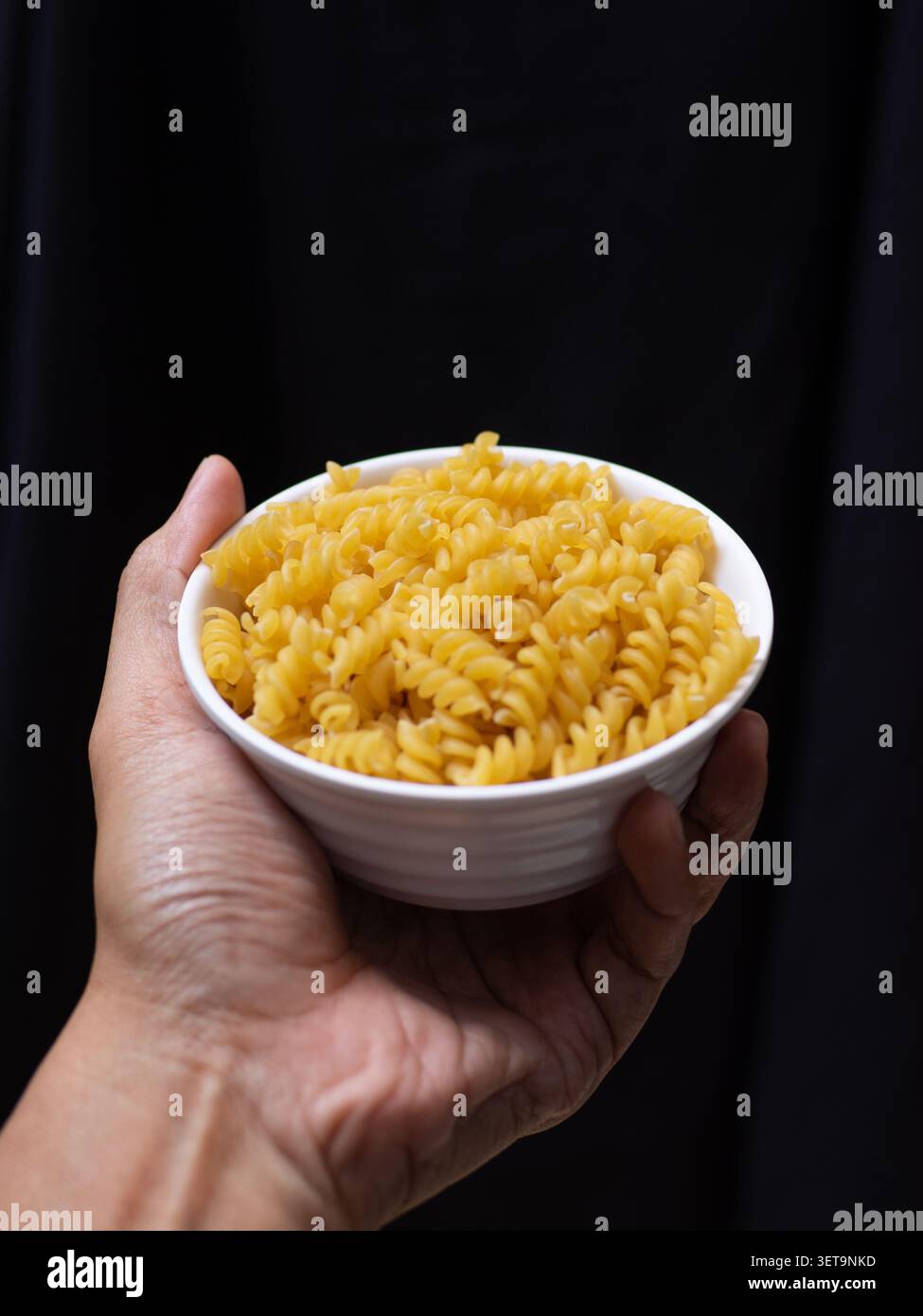 Fusilli secchi crudi su una ciotola bianca tenuta da una mano sinistra su sfondo nero Foto Stock