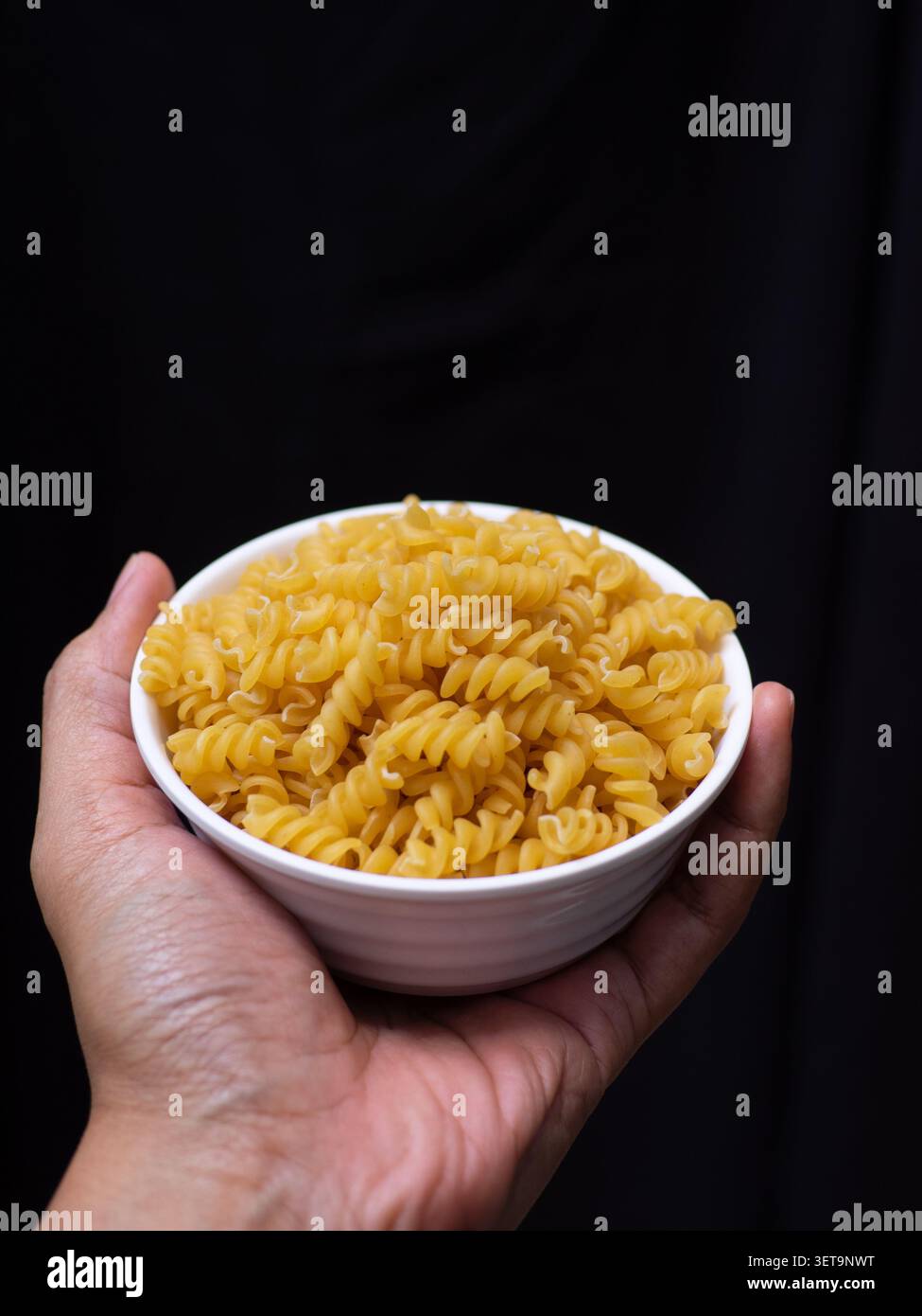 Fusilli secchi crudi su una ciotola bianca tenuta da una mano sinistra su sfondo nero Foto Stock
