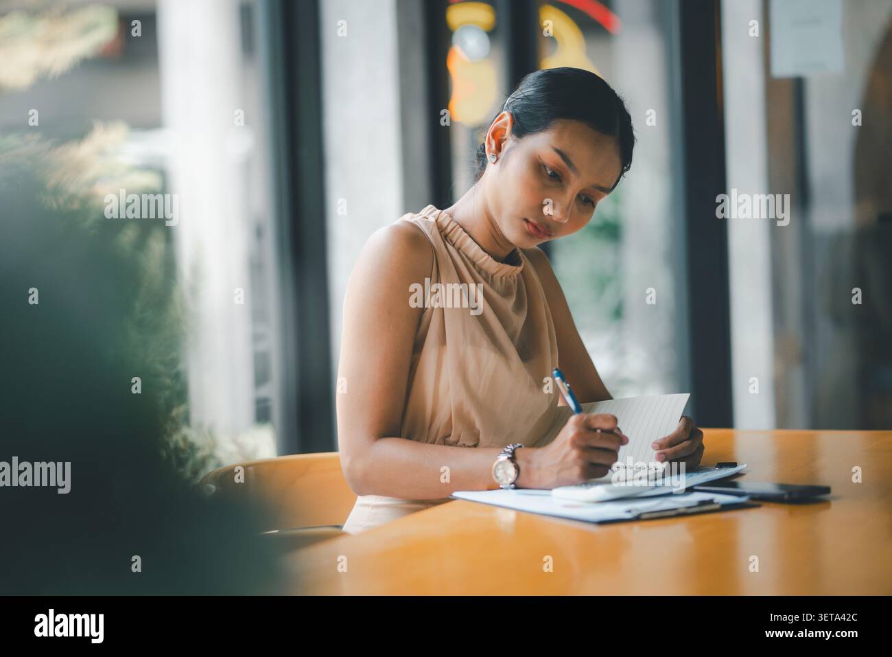 Donna d'affari asiatica che scrive appunti su un notebook nell'area di lavoro del bar con report finanziari, calcolatrice e smartphone, focalizzata su idee contabili, sbagli Foto Stock