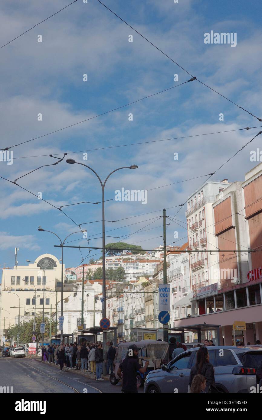 La strada urbana di Lisbona con cavi tranviari sopraelevati, lampioni stradali e pedoni contro il paesaggio urbano collinare Foto Stock
