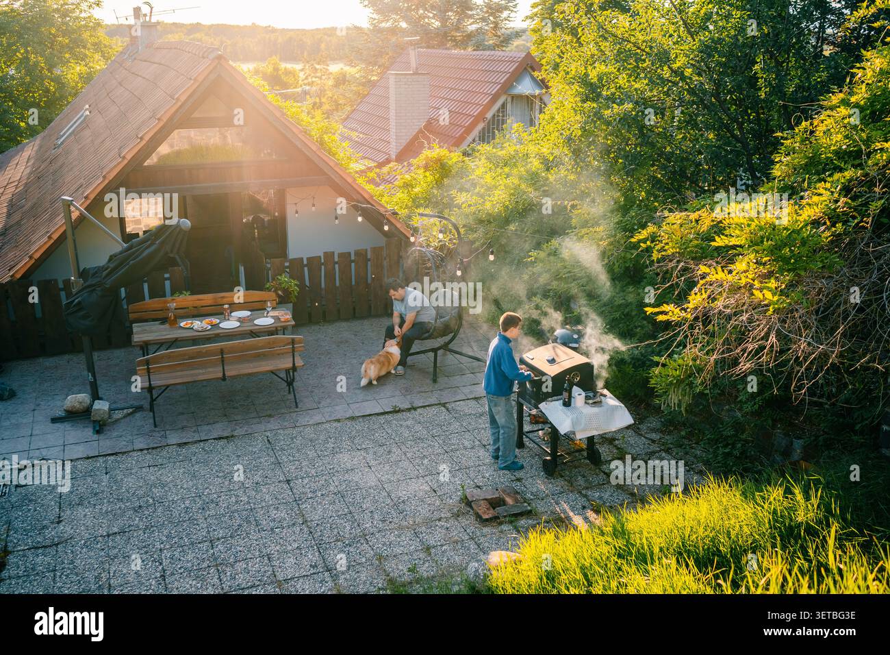 Famiglia che cucina al barbecue fuori da una casa al tramonto con un cane nelle vicinanze e un tavolo apparecchiato per un pasto Foto Stock