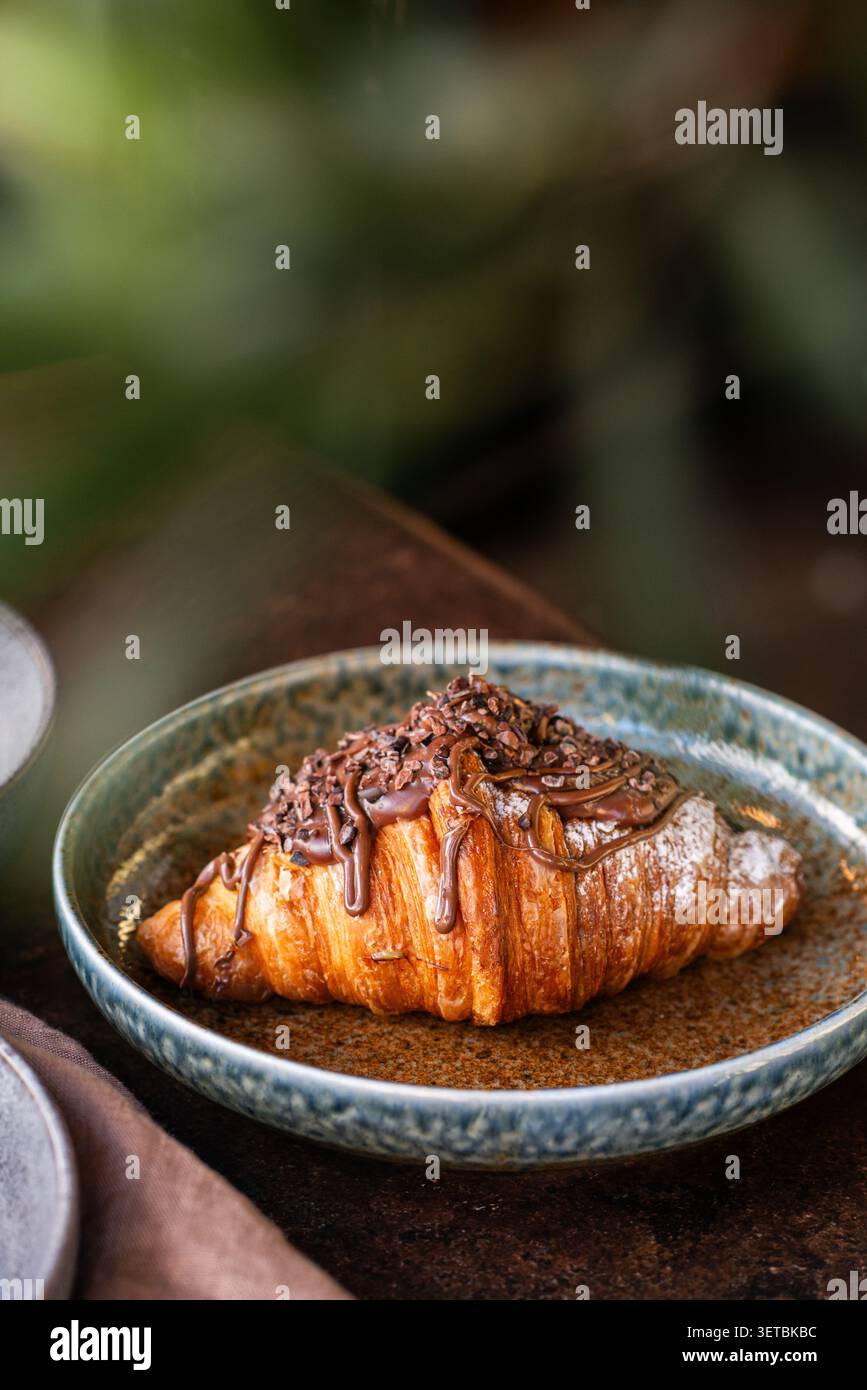 Un croissant di pasta sfoglia dorata condito con una ricca salsa di cioccolato e ricoperto di croccanti pennini di cacao, servito in una rustica ciotola di ceramica. Foto Stock