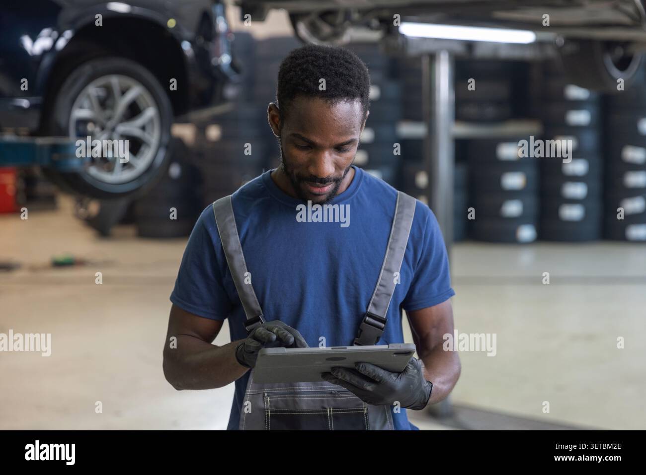 Meccanico afroamericano che utilizza un tablet per diagnosticare guasti, controllare la manutenzione e ordinare parti, accanto alle auto sollevate, ritratto. Foto Stock