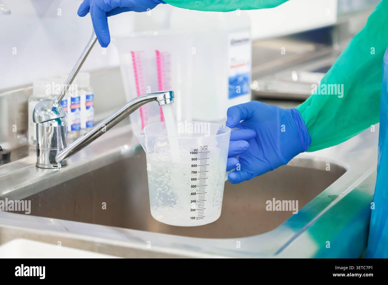 Un tecnico che indossa guanti in lattice blu riempie un contenitore graduato sotto un rubinetto per misurare con precisione i fluidi per la miscelazione chimica o l'igienizzazione Foto Stock