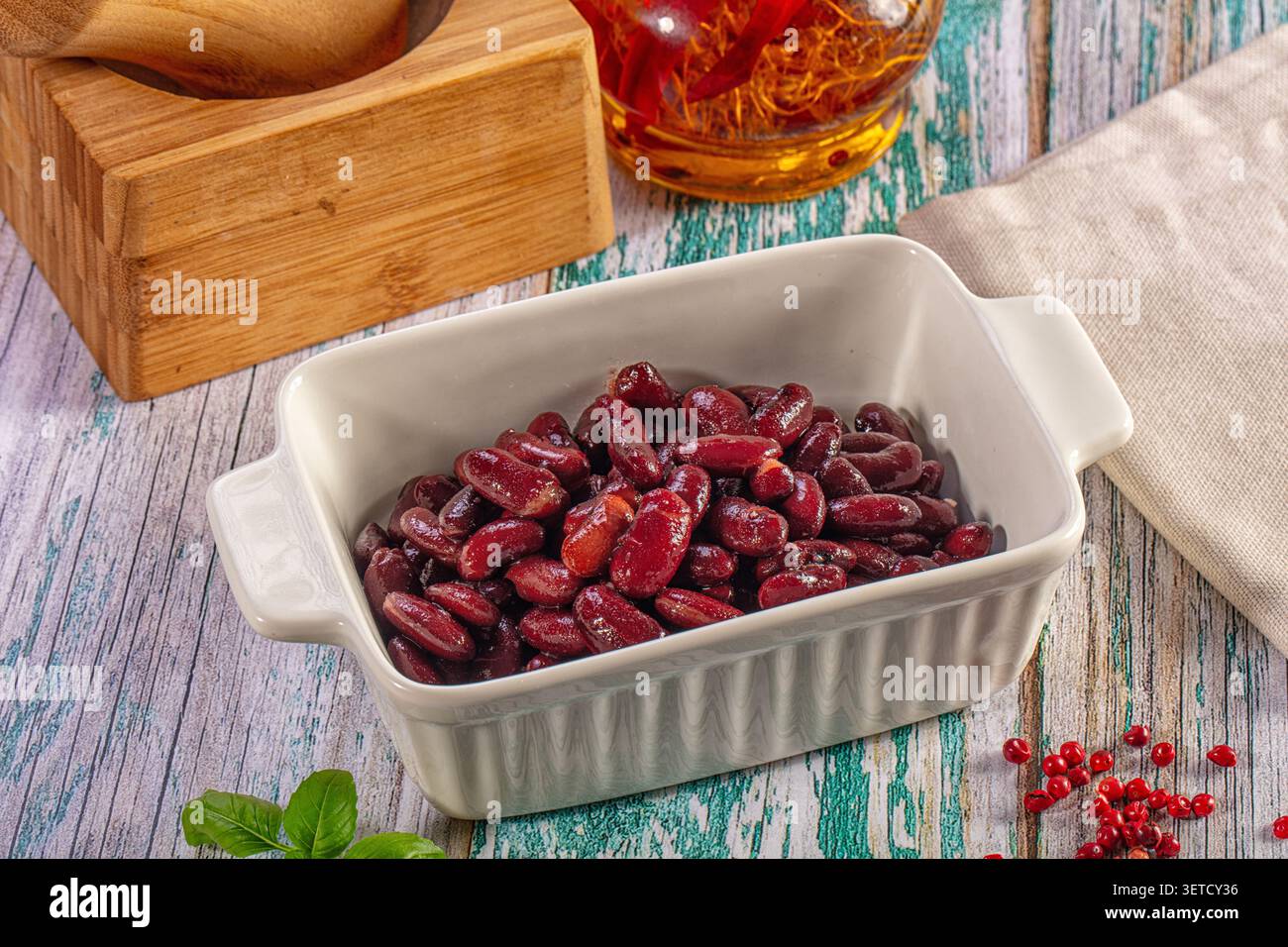 Primo piano di fagioli rossi bolliti in un piatto di ceramica bianca. La scena rustica include olio di peperoncino, spezie e basilico su un tavolo di legno in difficoltà. Guarire Foto Stock