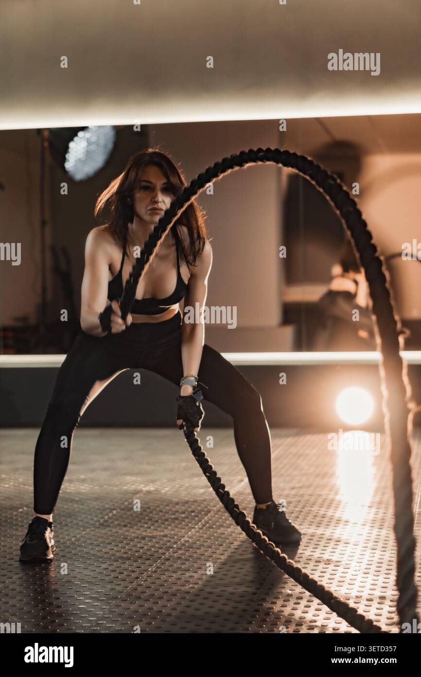Atletica femminile si allena con corde da battaglia, mostrando movimento potente e dedizione alla salute fisica. L'ambiente contemporaneo della palestra enfatizza lo stile di vita attivo e i risultati personali Foto Stock