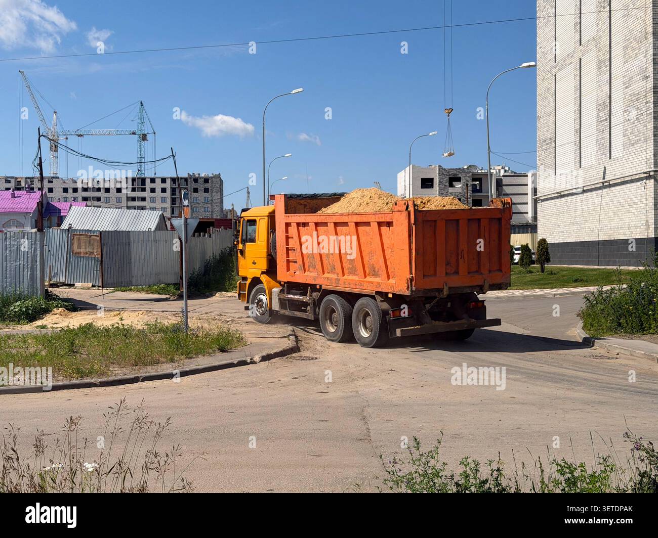 Un dumper arancione ha consegnato un carico di sabbia a un cantiere urbano con edifici residenziali e gru a torre. - Immagine stock catturata con smartphone