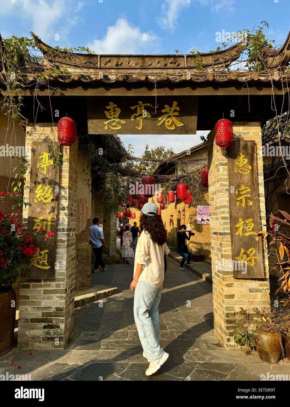 Donna che cammina attraverso l'arco decorato della porta del villaggio cinese tradizionale con lanterne rosse e calligrafia dorata, Guangxi, Cina - Immagine stock catturata con smartphone