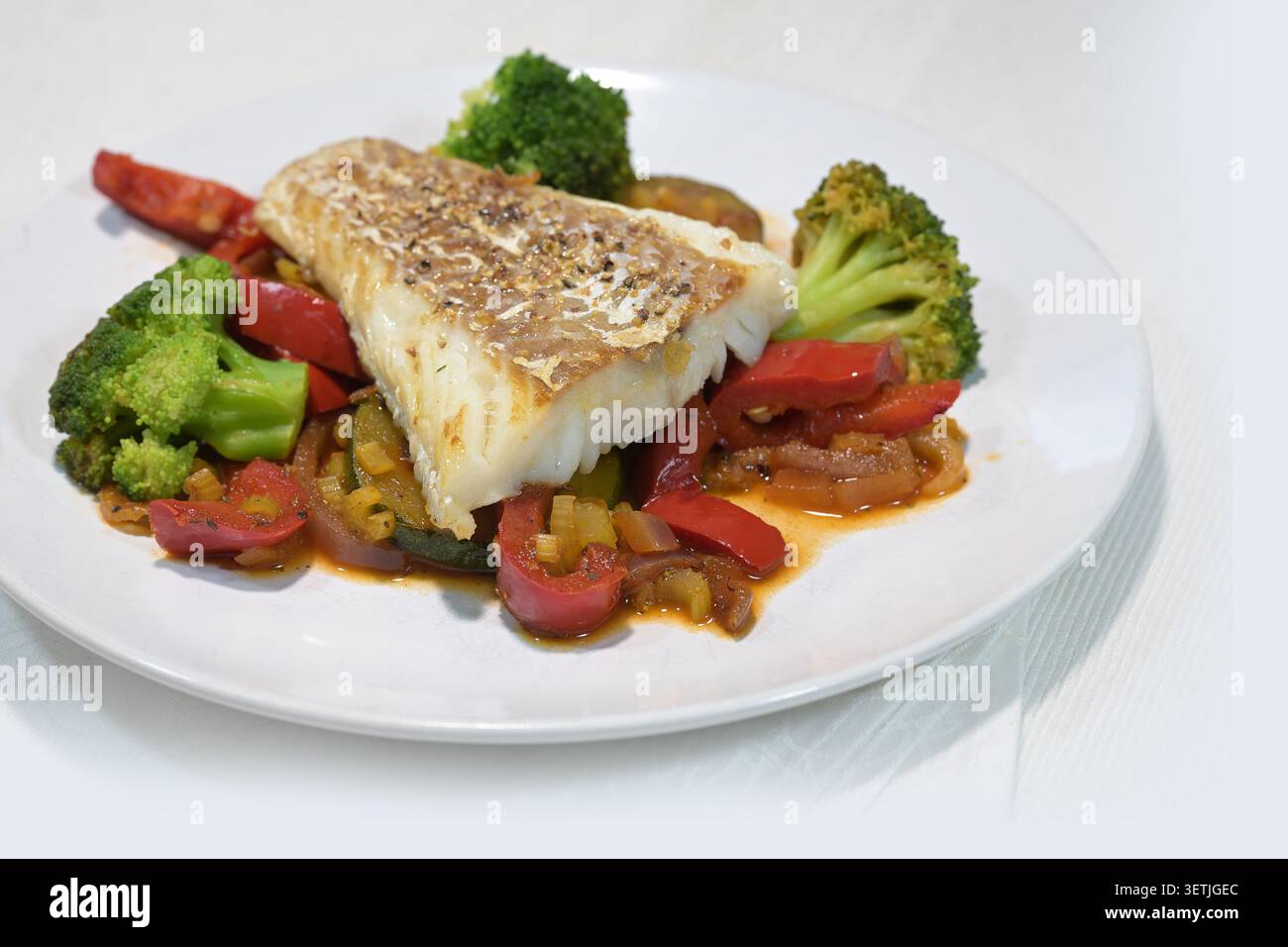 Filetto di merluzzo fritto su verdure come peperone rosso, broccoli e zucchine su un piatto bianco, piatto sano con proteine, vitamina e noci importanti Foto Stock