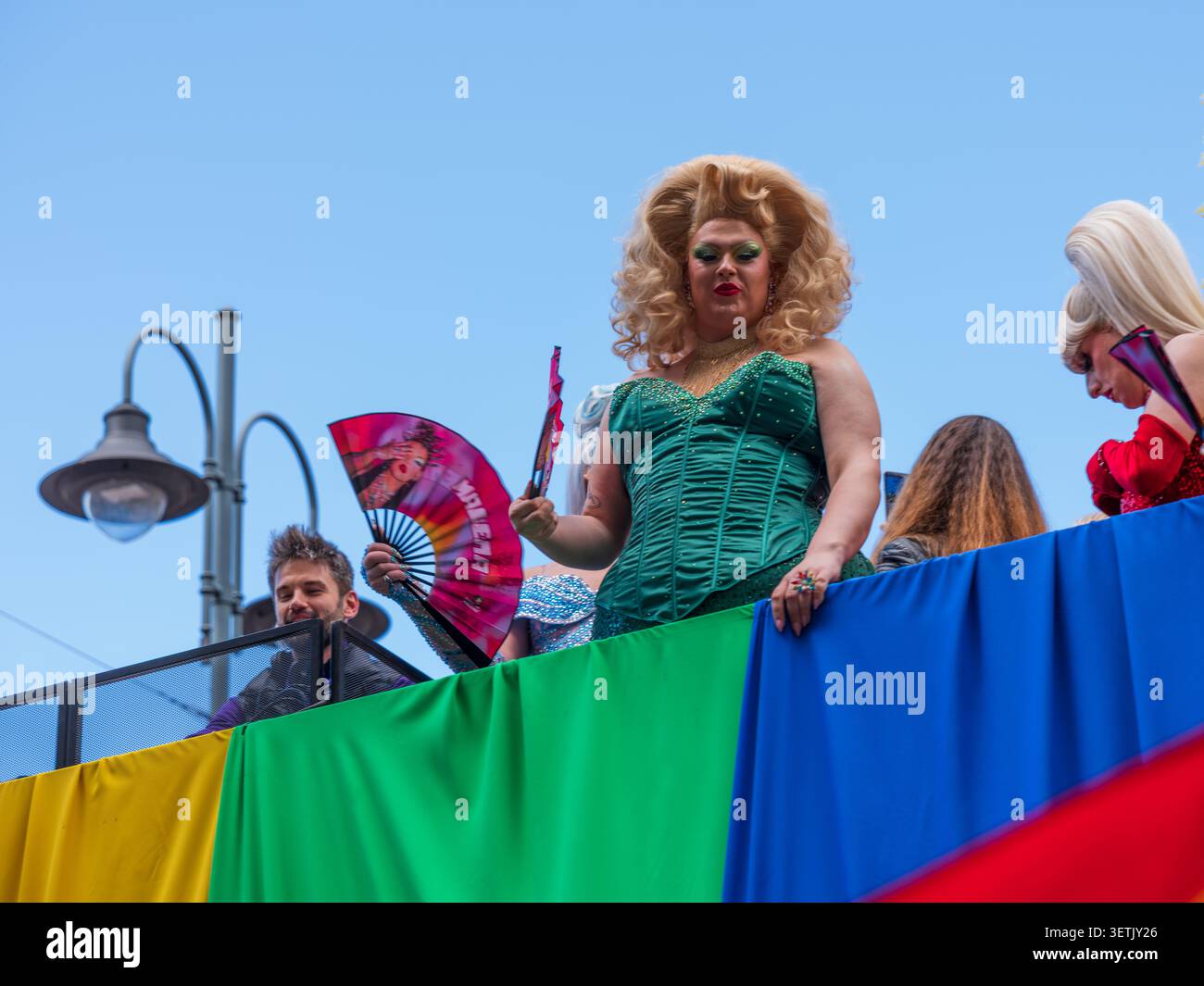 Vilnius, Lituania - 06 giugno 2026: Vista di una drag queen in un corsetto verde e parrucca bionda che tiene un ventilatore su una parata color arcobaleno galleggiante durante un prid Foto Stock