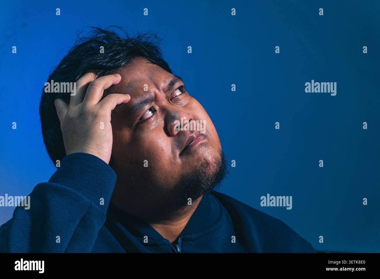Un soggetto maschile adulto stressato appoggia saldamente la mano contro la fronte sudata mentre guarda verso l'alto con un'espressione profondamente turbata, rivelando un'inte Foto Stock