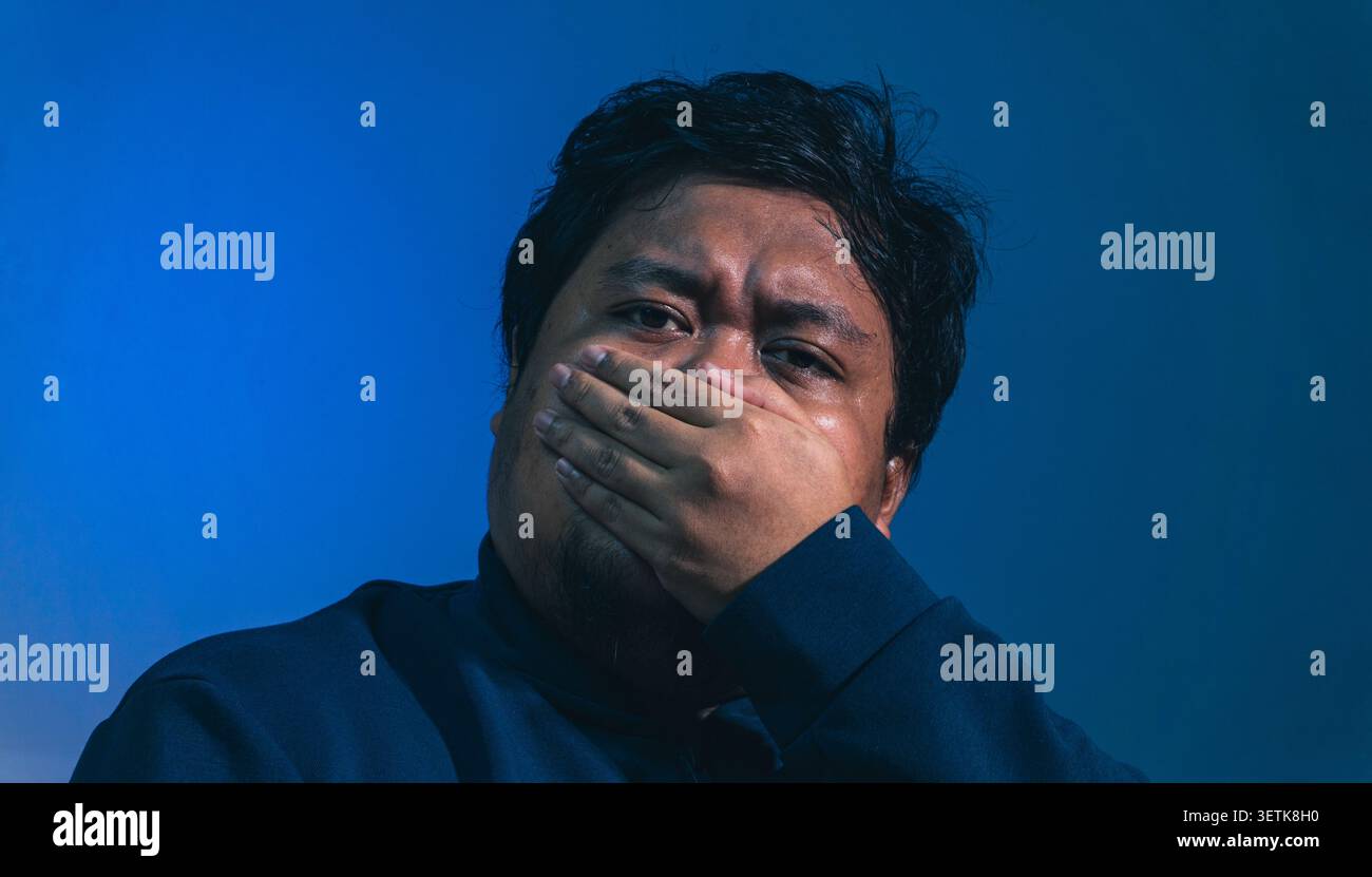 Un temibile soggetto maschile adulto fissa direttamente lo spettatore, coprendo saldamente la bocca con una mano, trasmettendo un senso di panico silenzioso, nascosto Foto Stock