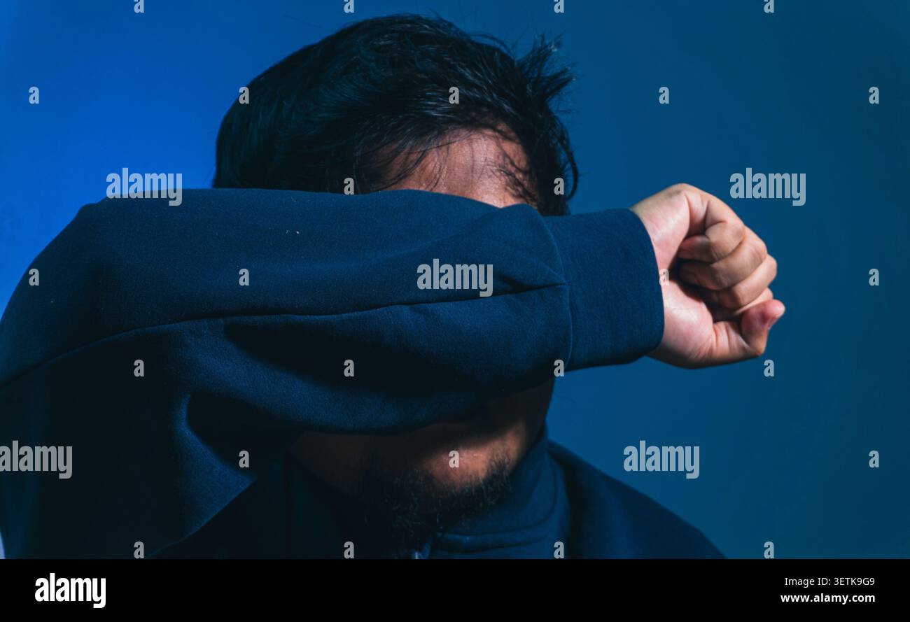 Un uomo difensivo alza la sua manica scura per nascondere completamente gli occhi e il viso, mostrando paura, protezione intensa o una profonda str emotiva Foto Stock