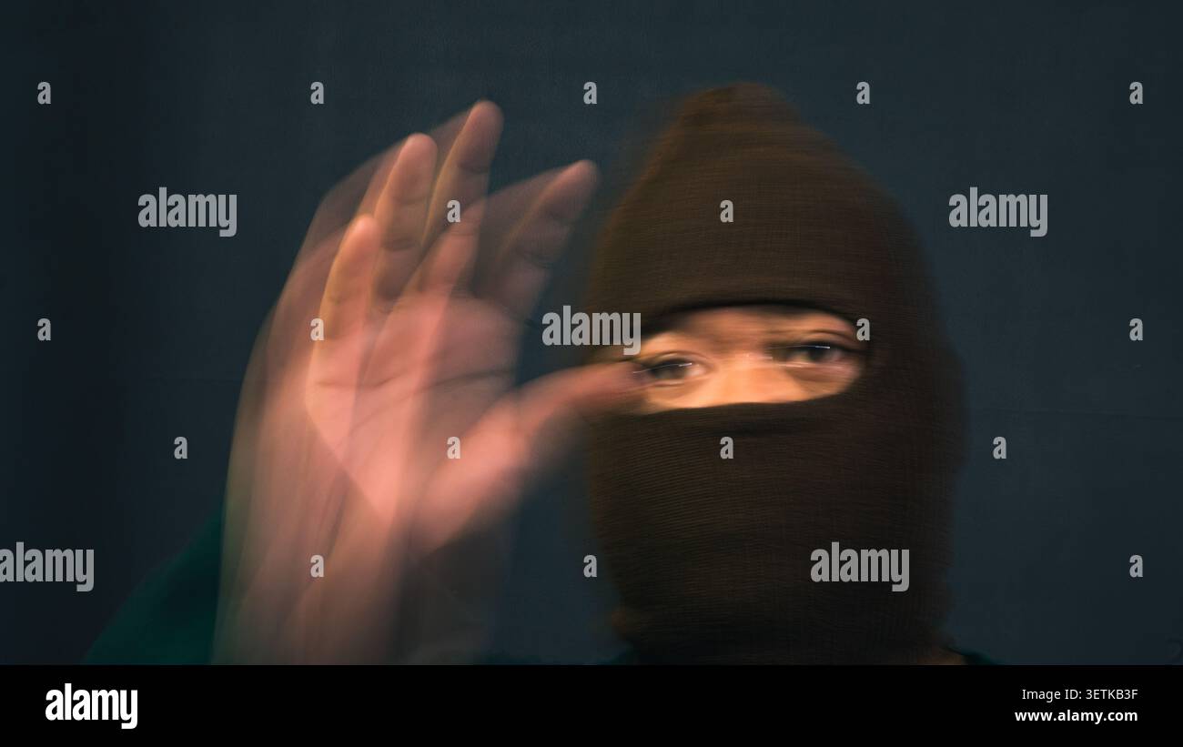Un uomo che indossa una balaclava a maglia marrone scuro guarda avanti mentre alza la mano sinistra con un movimento sfocato che crea sentieri spettrali sul viso Foto Stock