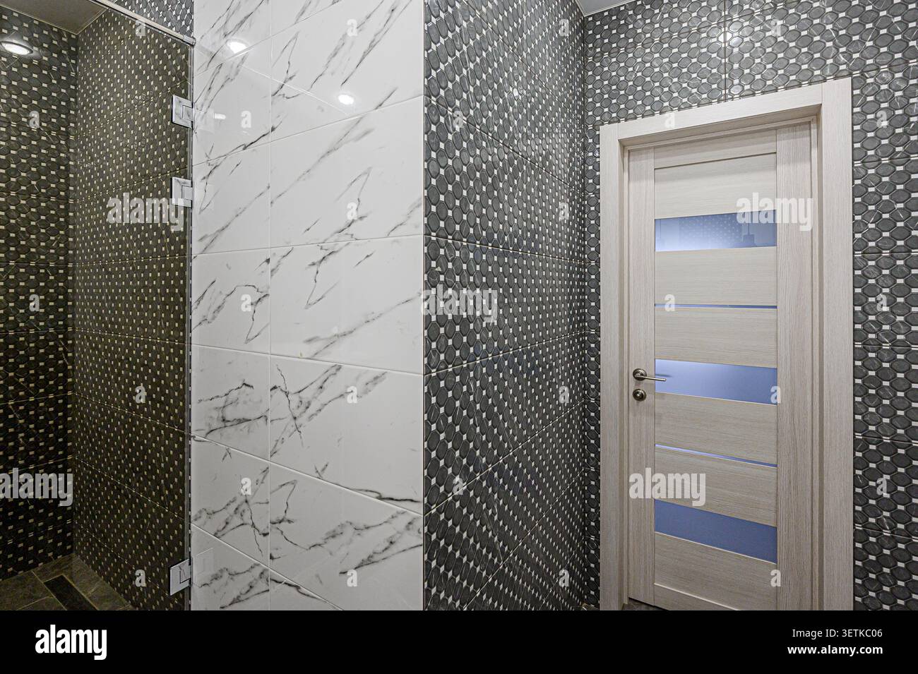 Un bagno moderno caratterizzato da pareti piastrellate in motivi geometrici neri e marmo bianco, con una porta in legno chiaro con accenti in vetro Foto Stock