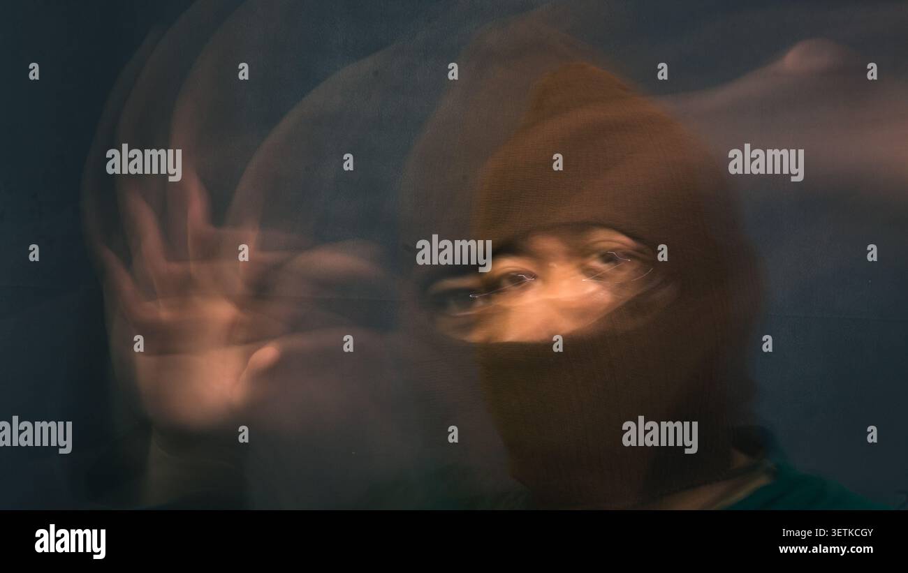 Una figura maschile in un balaclava marrone lavorato a maglia alza la mano con un movimento estremamente sfocato verso l'alto formando percorsi fantasma traslucidi e curvi sulla sinistra. fa Foto Stock