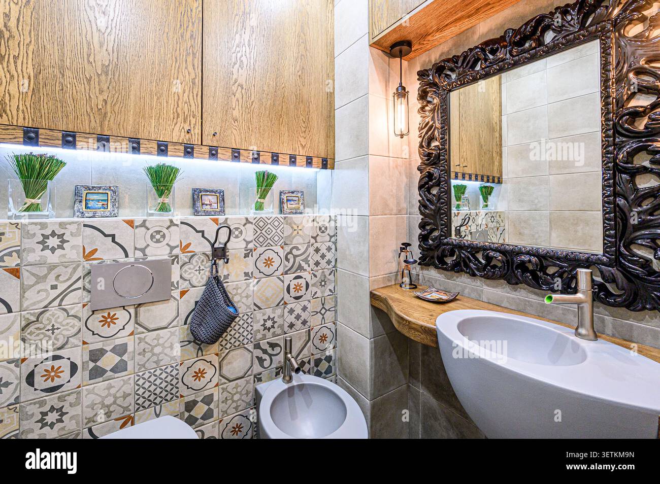 Bagno elegante con specchio decorato, piastrelle a motivi geometrici e accenti in legno. Un design moderno e unico che fonde elementi rustici e contemporanei. Foto Stock