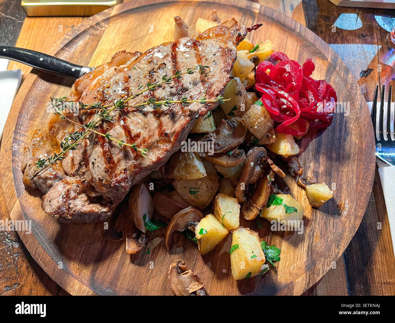 Primo piano ad angolo alto che mostra bistecca di carne grigliata con timo, servita con patate arrosto, funghi e cipolla rossa sottaceto su tavola rotonda di legno Foto Stock
