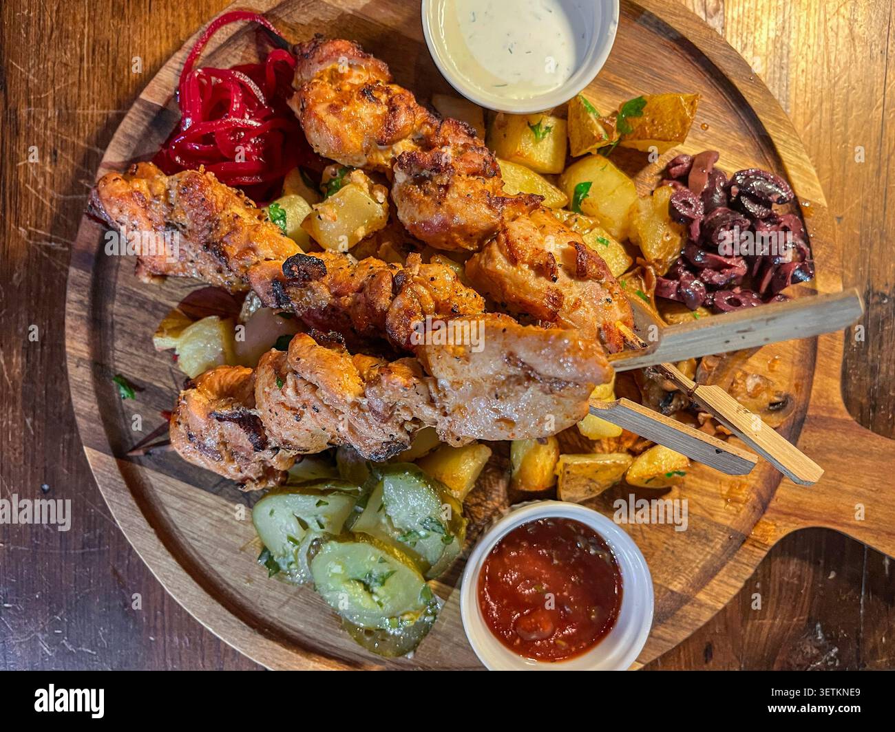 Tagliere in legno con spiedini di carne alla griglia, patate arrosto, cetrioli sottaceto, cipolle rosse, olive, e salse da immersione, creando un rustico sostanzioso Foto Stock
