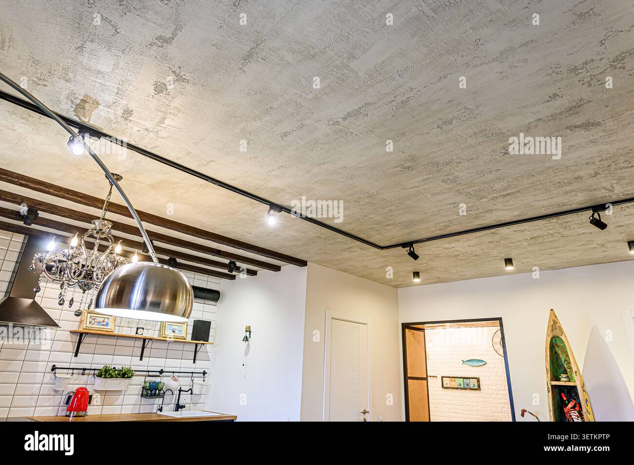 Interni moderni con soffitto testurizzato, illuminazione binari e una combinazione di elementi industriali e classici. Angolo cucina visibile con lampadario Foto Stock