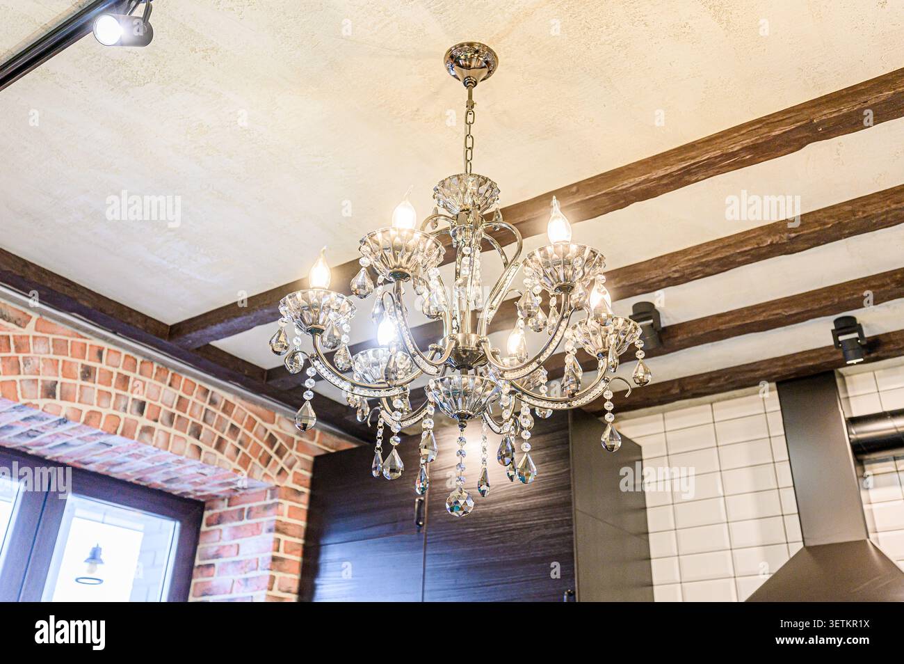 Un classico lampadario di cristallo pende da un soffitto con travi in legno. Sono visibili anche mattoni e piastrelle bianche Foto Stock