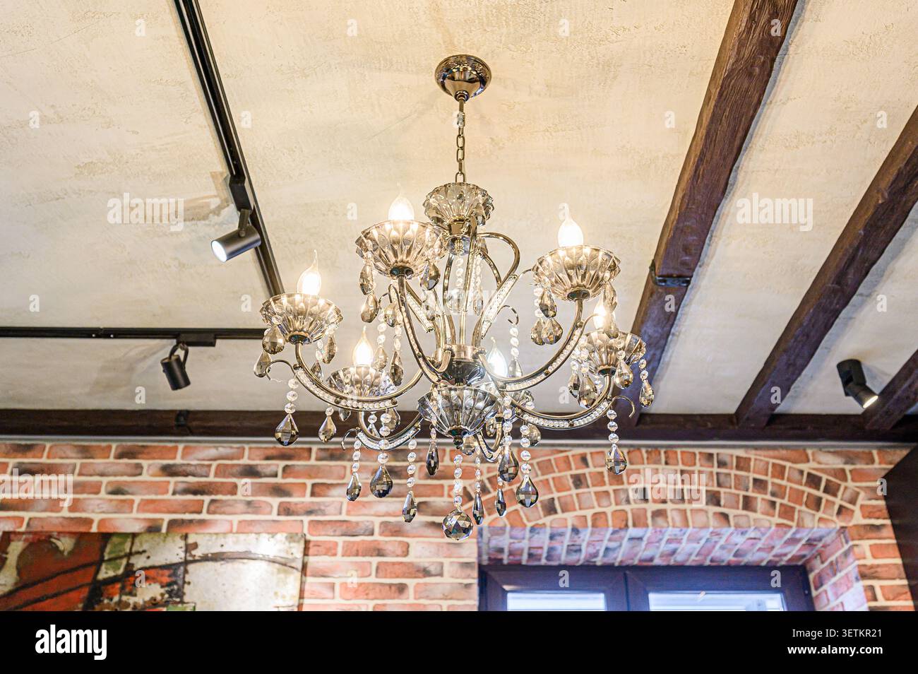 Un lampadario di cristallo è appeso sotto un soffitto con travi in legno a vista e illuminazione a binario. La muratura è visibile nella parte inferiore del telaio Foto Stock