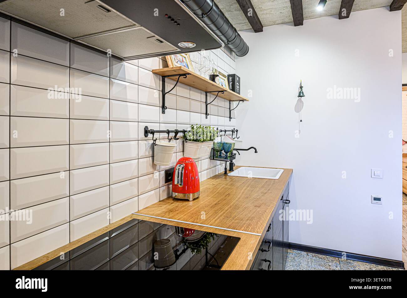 Una cucina moderna caratterizzata da pareti piastrellate bianche, un ripiano in legno e accenti neri. Un bollitore rosso aggiunge un tocco di colore. Gli scaffali contengono piante e decorazioni Foto Stock