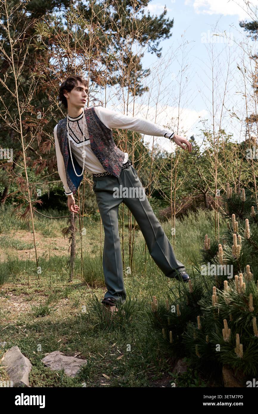 Un giovane uomo con un gilet a motivi geometrici e una blusa in pizzo pone con sicurezza la natura Foto Stock