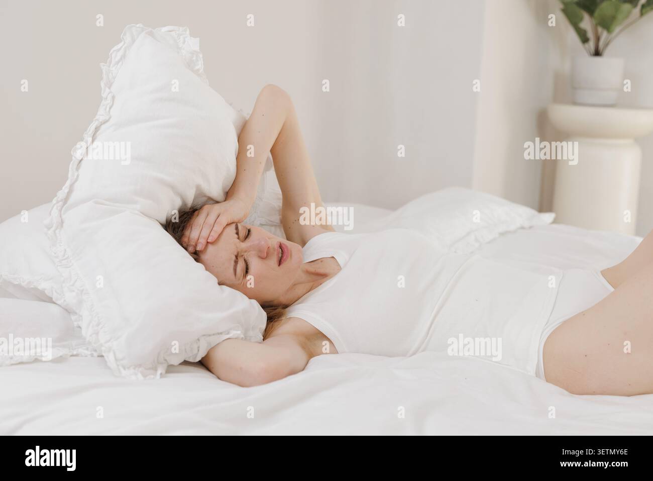 Una donna è sdraiata a letto, visibilmente angosciata a causa di un grave mal di testa, evidenziando i problemi di salute delle donne e l'impatto dell'emicrania. Foto Stock