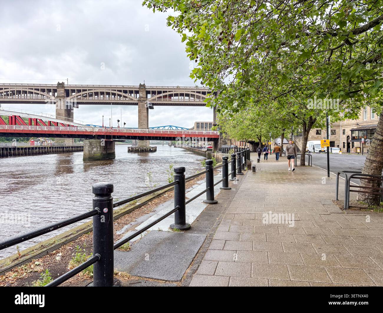 Persone che camminano su un sentiero lastricato lungo il fiume Tyne, caratterizzato dal ponte Tyne e dall'architettura industriale storica di Newcastle upon Tyne Foto Stock