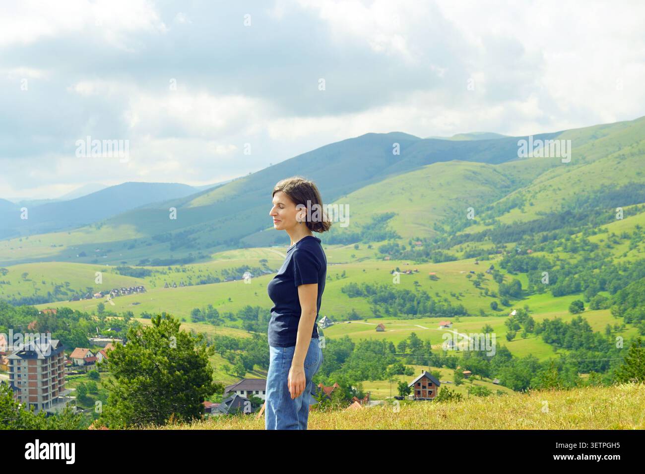 La visitatrice si erge sopra la valle stratificata e le creste verdi. Mostra un rifugio benessere, un'esperienza di fuga tranquilla in montagna e prospettive di viaggio lento. Foto Stock