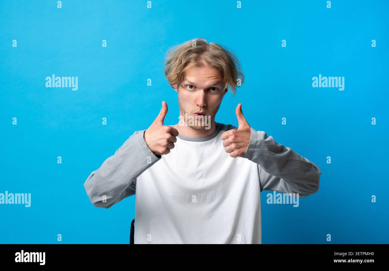 Giovane biondo che fa un doppio gesto, approva un servizio eccellente, mostra accordo, positività, successo, e pieno consenso su blue Foto Stock